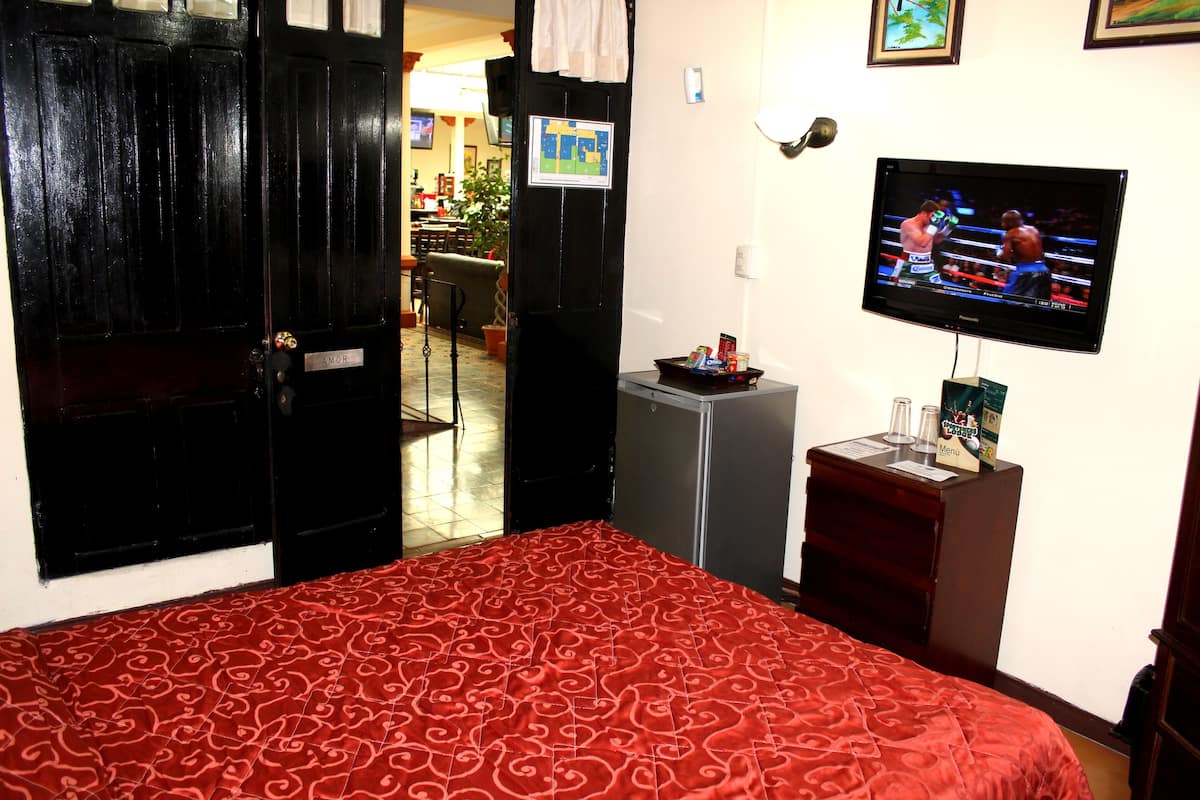 Een hotelkamer met een rood bed met een decoratieve spreidek, een aan de muur bevestigde televisie, een minibars en een deur met een naambordje.