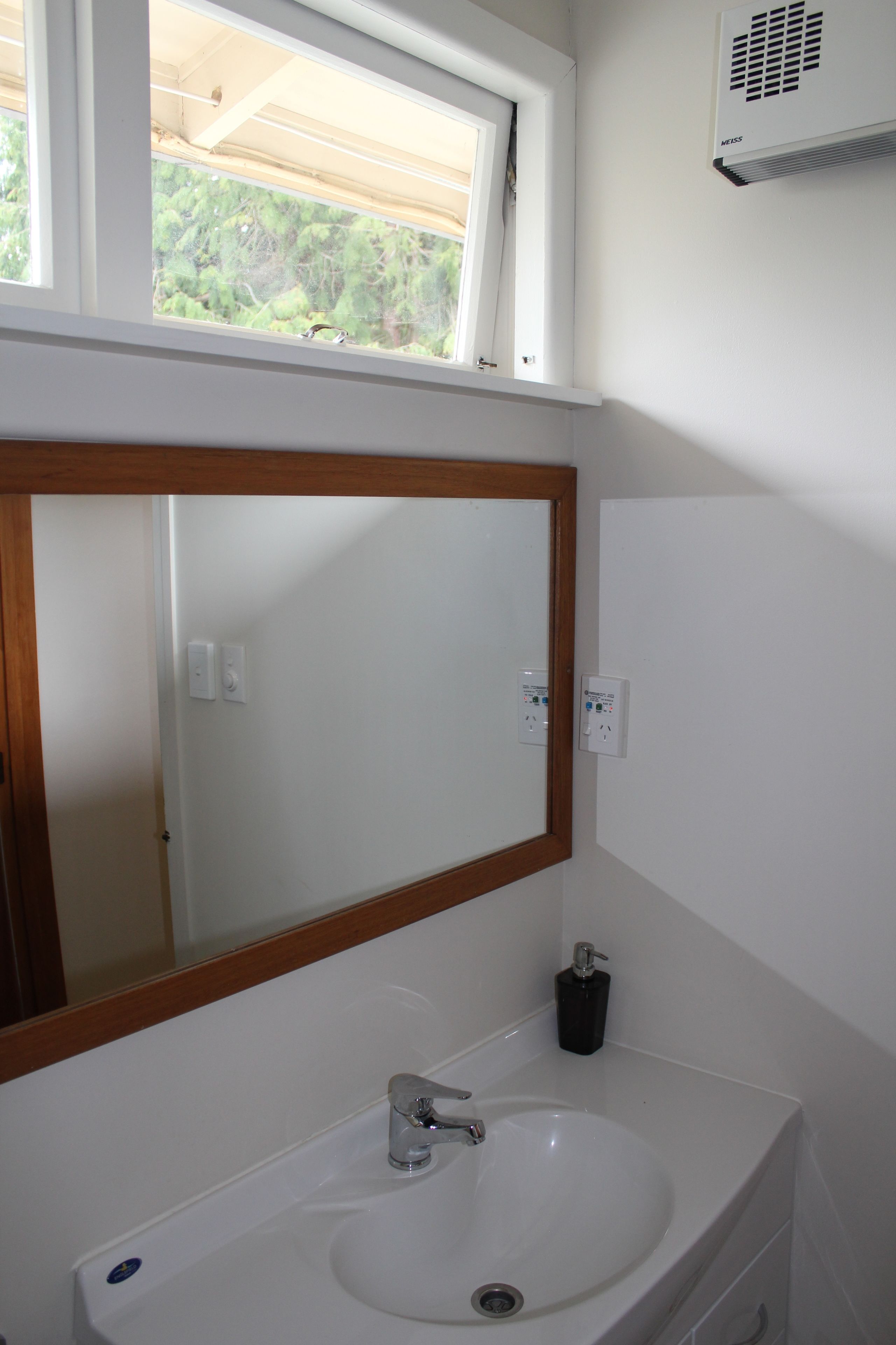 A bathroom with a white sink, a mirror with a wooden frame, a window above the sink, and a wall-mounted air conditioner.