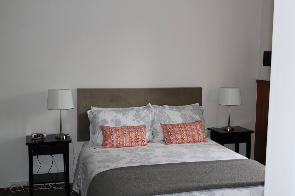 Una cama bien hecha con almohadas estampadas, dos mesitas de noche con lámparas y un reloj de pared.