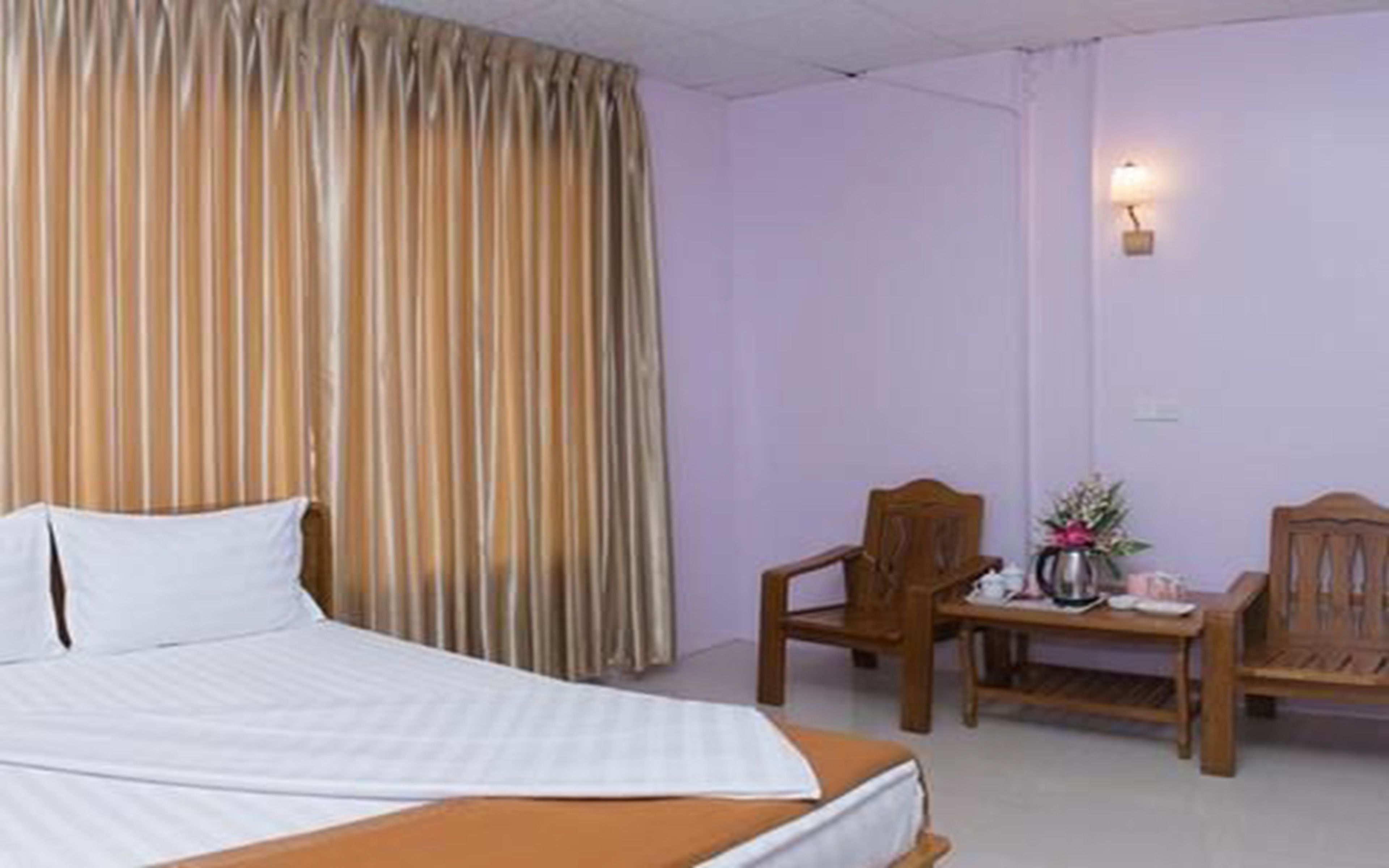 A hotel room with a bed, a wooden chair, a small table with a vase of flowers, and a wall-mounted lamp.