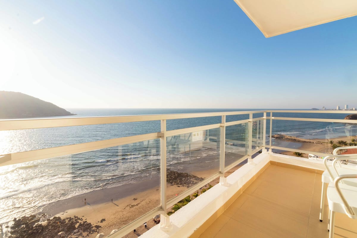 A balcony with a clear view of the beach and ocean, featuring a white railing and a few chairs.
