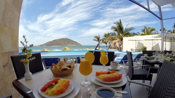 A table set with breakfast including fruit, juice, and coffee, overlooking a pool and beach. - Pacific Palace Beach Tower Hotel (Mazatlán)