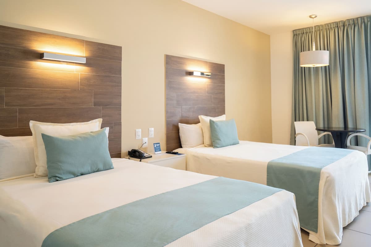 A hotel room with two beds, a desk, a chair, and a lamp.