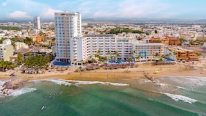 Un hôtel situé sur la côte, avec plusieurs étages, une plage équipée de parasols et de chaises longues, et une vue sur le paysage urbain environnant.