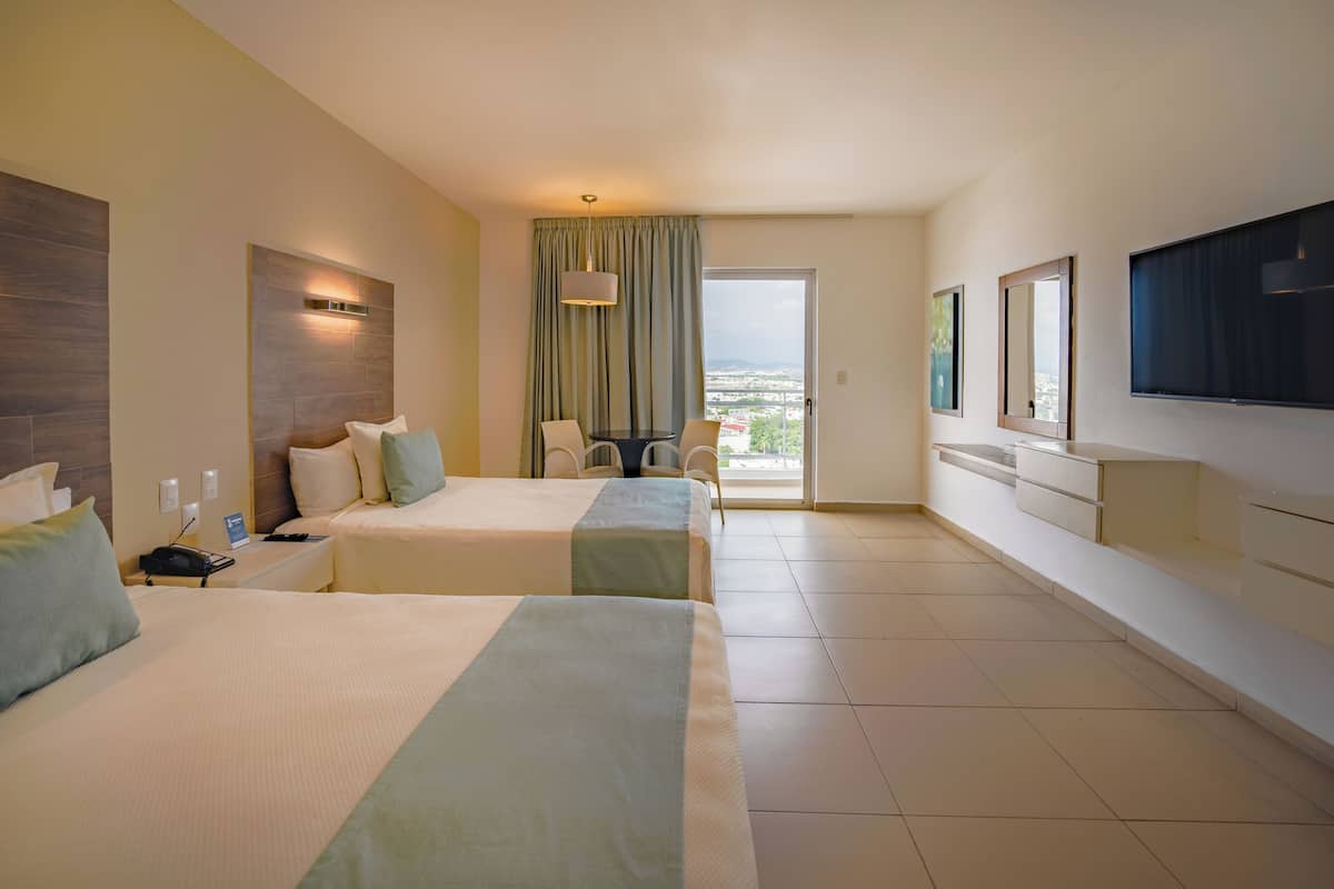 A hotel room with two beds, a dining table, chairs, a TV, and a balcony with a view.