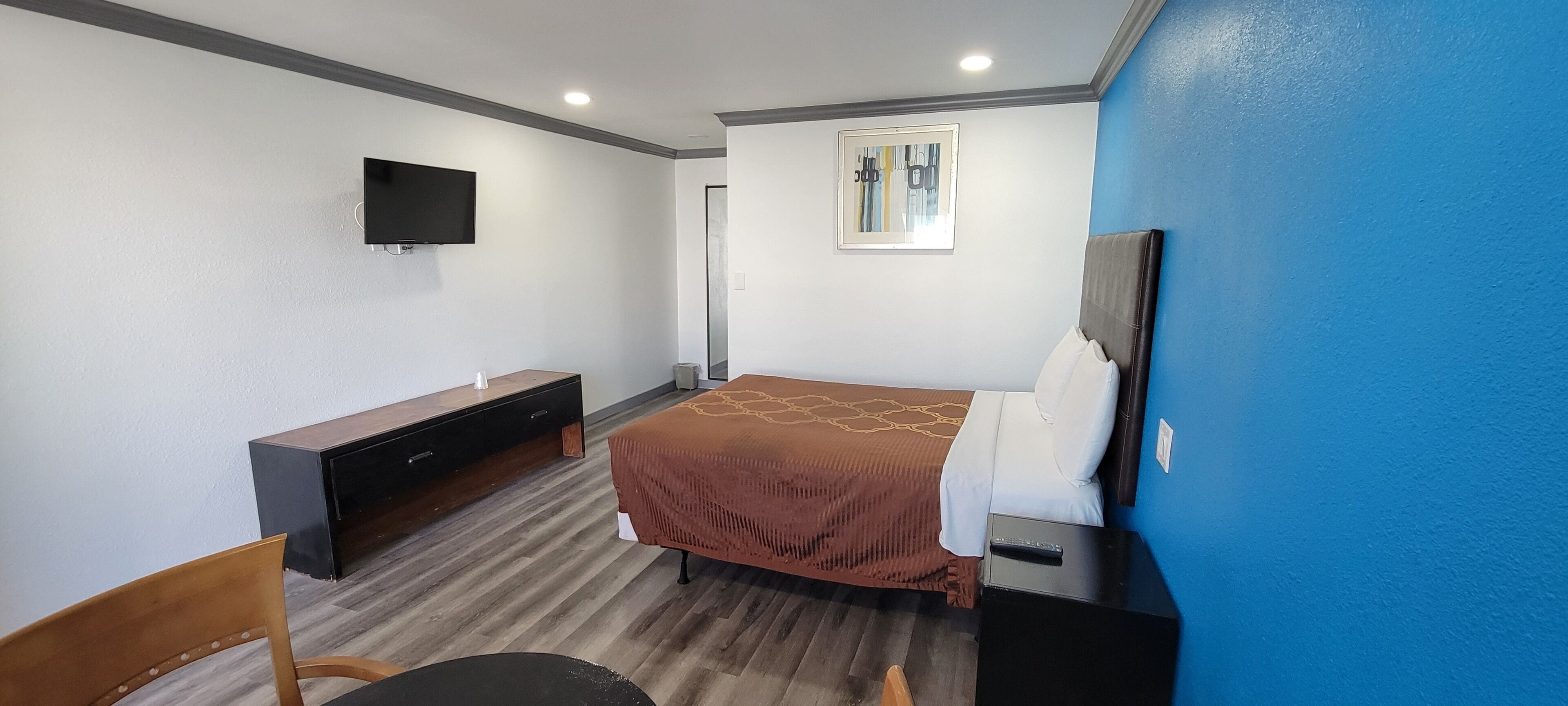 A bedroom with a bed, a TV mounted on the wall, a desk with a chair, and a window with blinds.