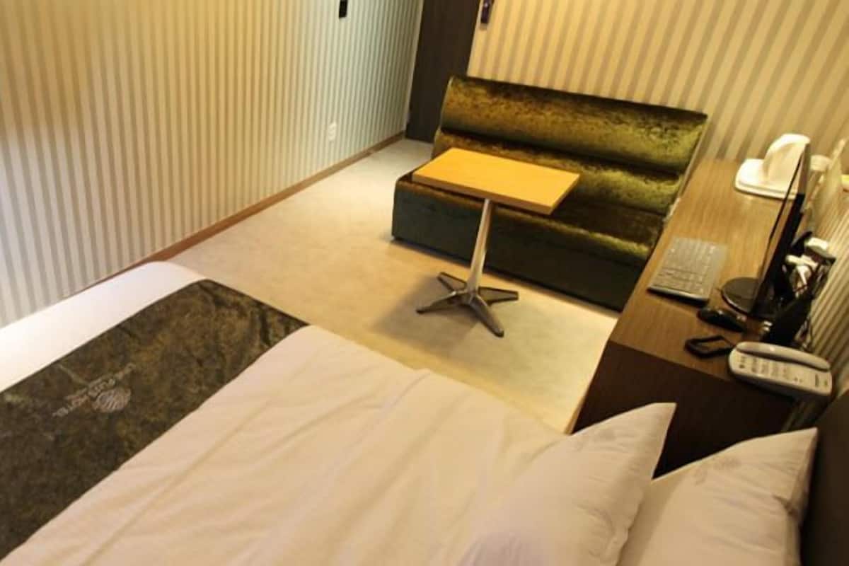 Quarto de hotel com cama, televisão na parede, sofá, mesa pequena e mesa de cabeceira com telefone e abajur.