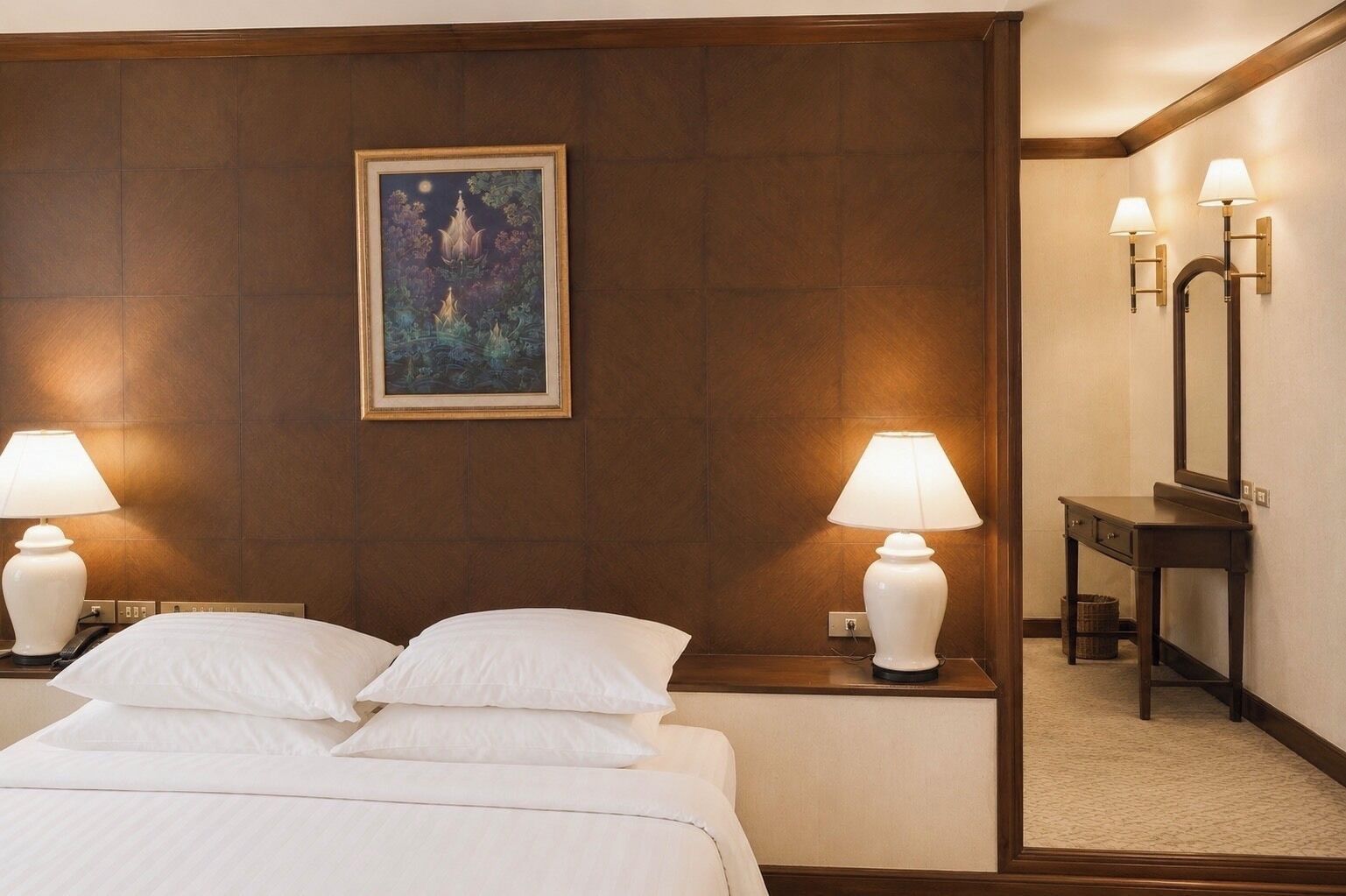 A hotel room with a bed, two bedside lamps, a nightstand, a mirror, and a framed painting on the wall.