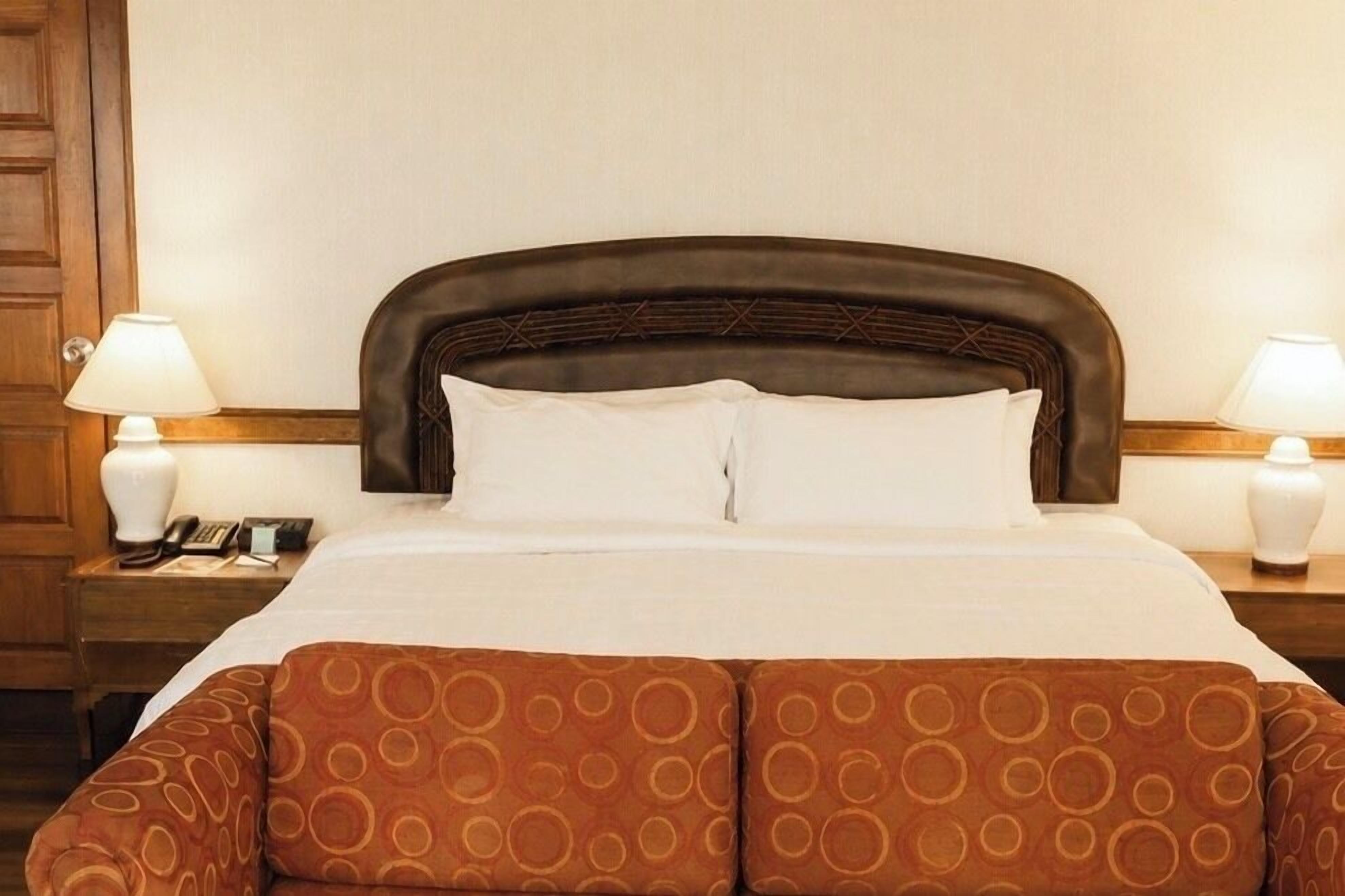 A hotel room with a large bed, two bedside lamps, a wooden nightstand, and a patterned throw pillow.