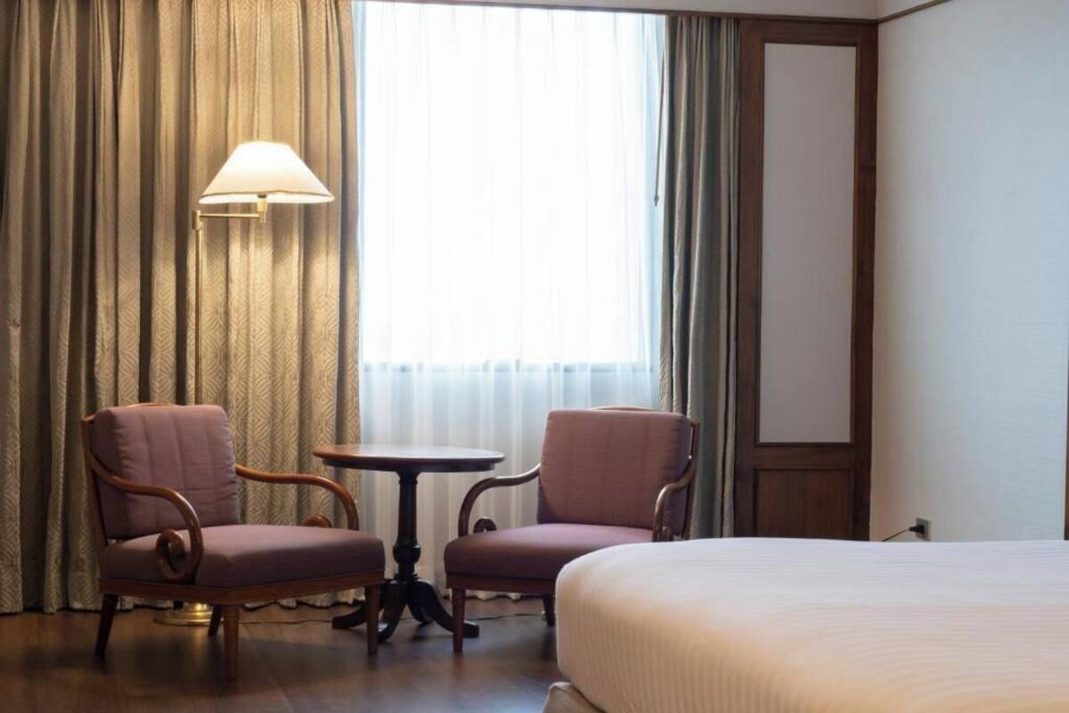 Habitación de hotel con una cama, dos sillones, una mesita, una lámpara y un amplio ventanal con cortinas.