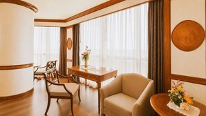 Ein Hotelzimmer mit Schreibtisch aus Holz, zwei Stühlen, einer Sitzecke und großen Fenstern mit Vorhängen.