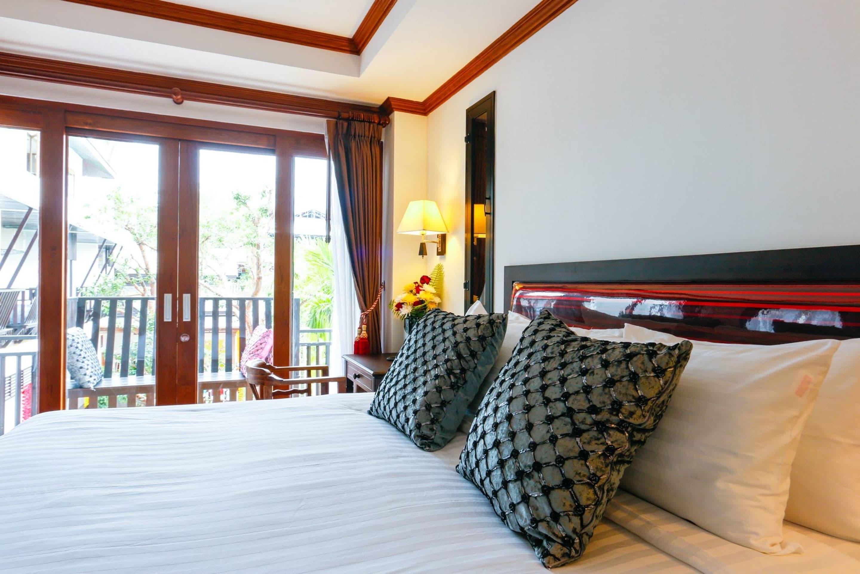 Ein Hotelzimmer mit einem großen Bett, zwei gemusterten Kissen, einem Nachttisch mit Lampe und Blick ins Freie durch eine Schiebetür.