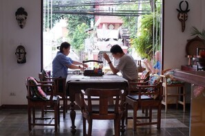A dining area with a wooden table and chairs, a view of a street with buildings and trees, and decorative wall art. - Baan Huen Phen (Chiang Mai)