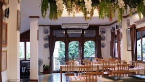 Un espace repas avec des tables et des chaises en bois, un plafond décoratif agrémenté de plantes suspendues et une vue sur l’extérieur grâce à de grandes fenêtres.