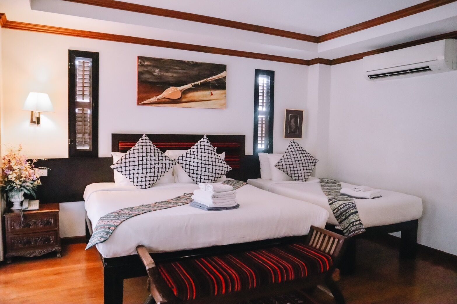 A hotel room with two beds, a wooden bench, a nightstand with a lamp, and a painting on the wall.