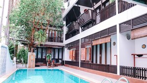 Une piscine avec une terrasse en bois, un espace salon couvert et un immeuble à plusieurs étages doté de balcons.