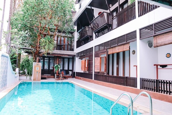 A swimming pool with a wooden deck, a covered seating area, and a multi-story building with balconies. - Baan Huen Phen (Chiang Mai)