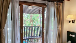 Une chambre d’hôtel avec une porte en bois donnant sur un balcon d’où l’on peut admirer les arbres et la piscine. La chambre est équipée d’un lit recouvert d’une couette blanche, d’une table de chevet avec une lampe et un téléphone.