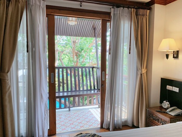 A hotel room with a wooden door leading to a balcony with a view of trees and a pool. There is a bed with a white comforter and a bedside table with a lamp and a telephone. - Baan Huen Phen (Chiang Mai)