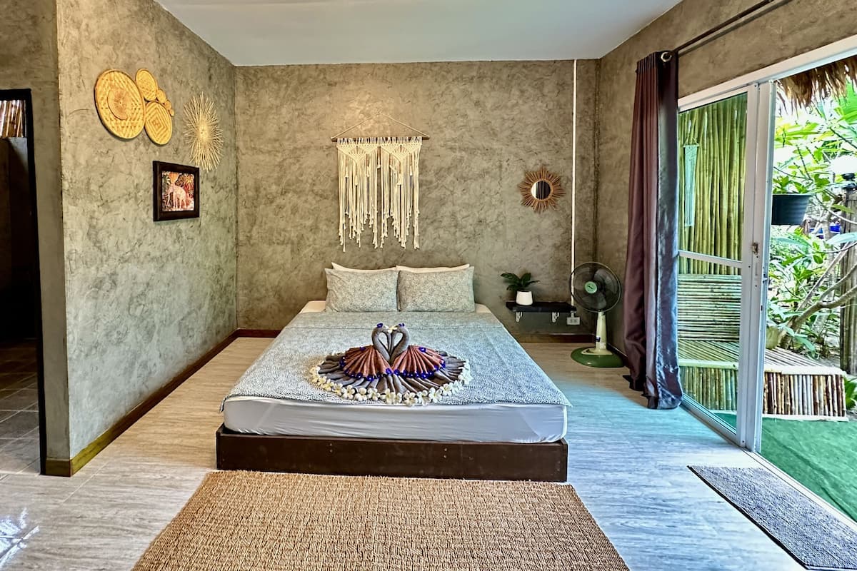A bedroom with a bed, a large window, a macrame wall hanging, and a picture frame.