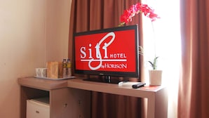 Habitación de hotel con una televisión mostrando la marca 'Siti Hotel by Horizon', un escritorio de madera con un control remoto, una pequeña planta en maceta y una botella de agua.