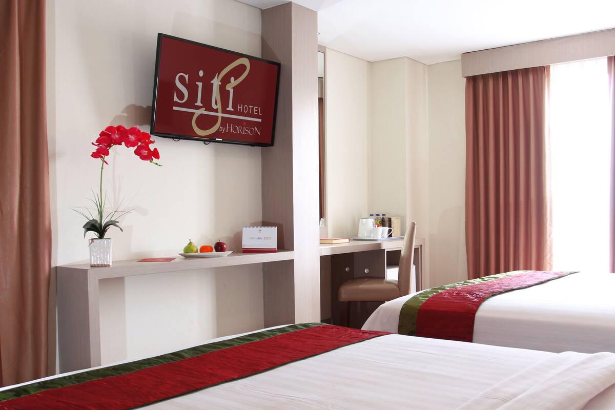 A hotel room with two beds, a desk, a TV displaying the hotel name, and a window with curtains.