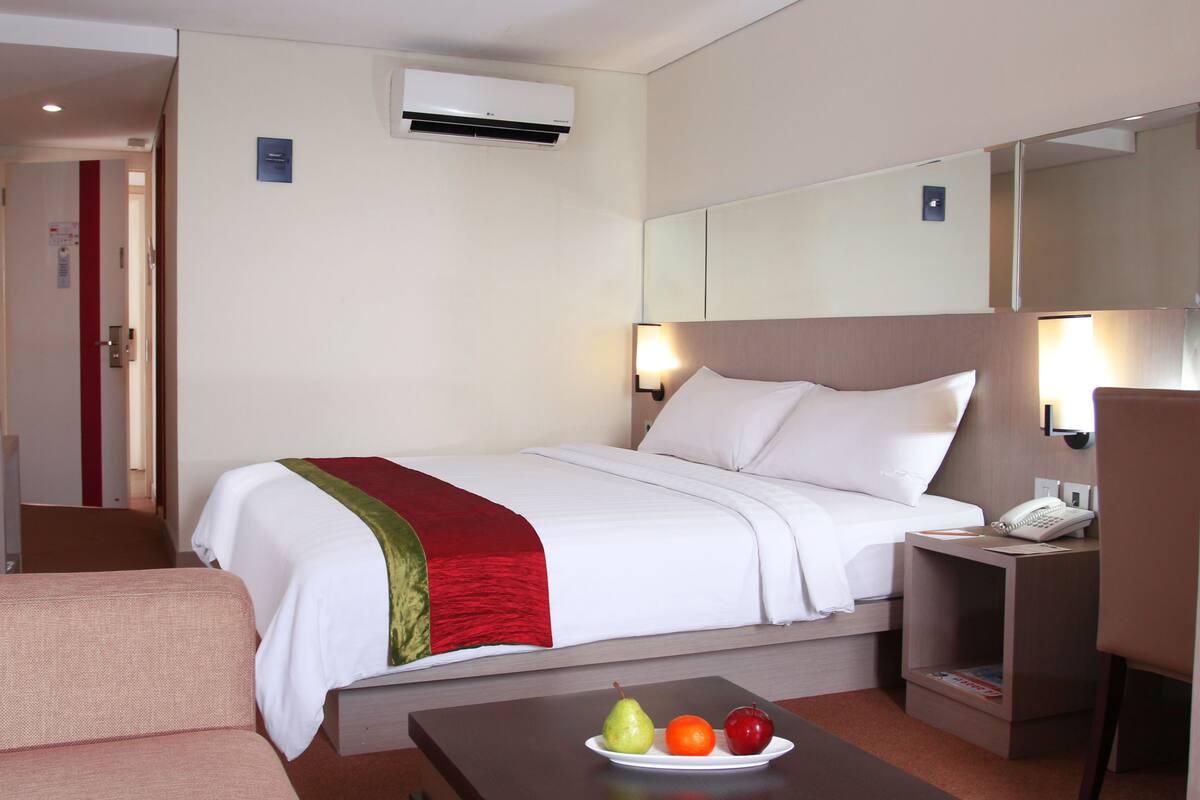 A hotel room with a bed, bedside table, chair, desk, and a small table with fruits.