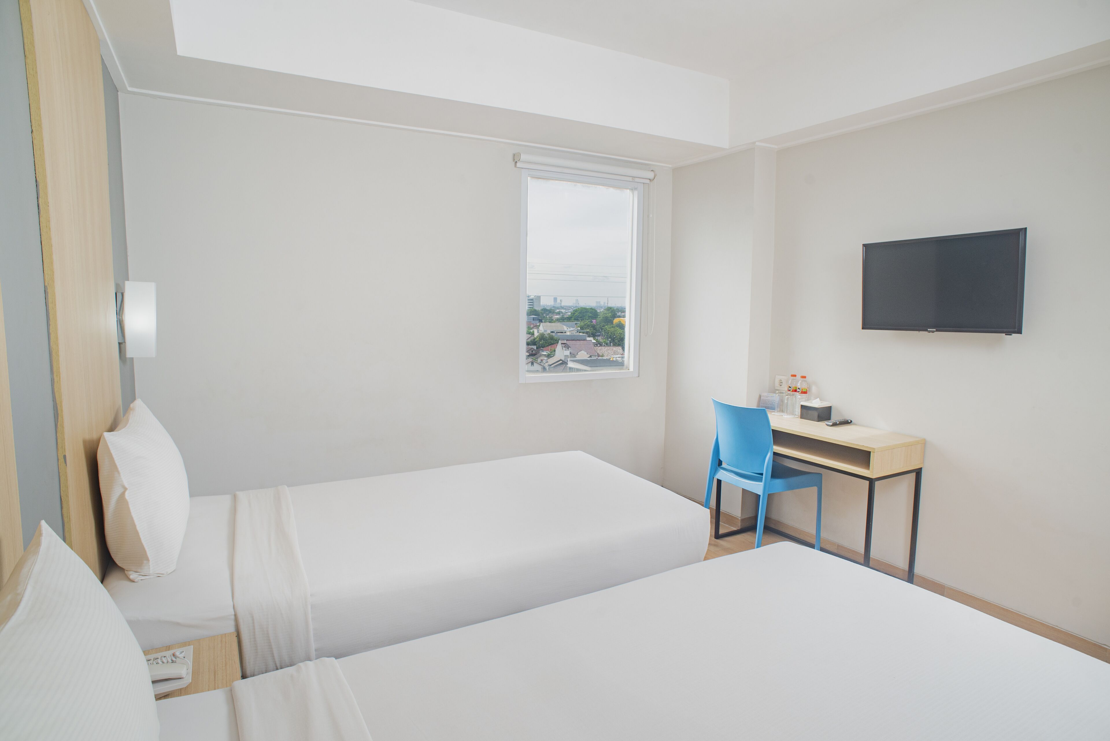 Una camera d'albergo con due letti singoli, una scrivania con una sedia, una TV a parete e una finestra che si affaccia su edifici.