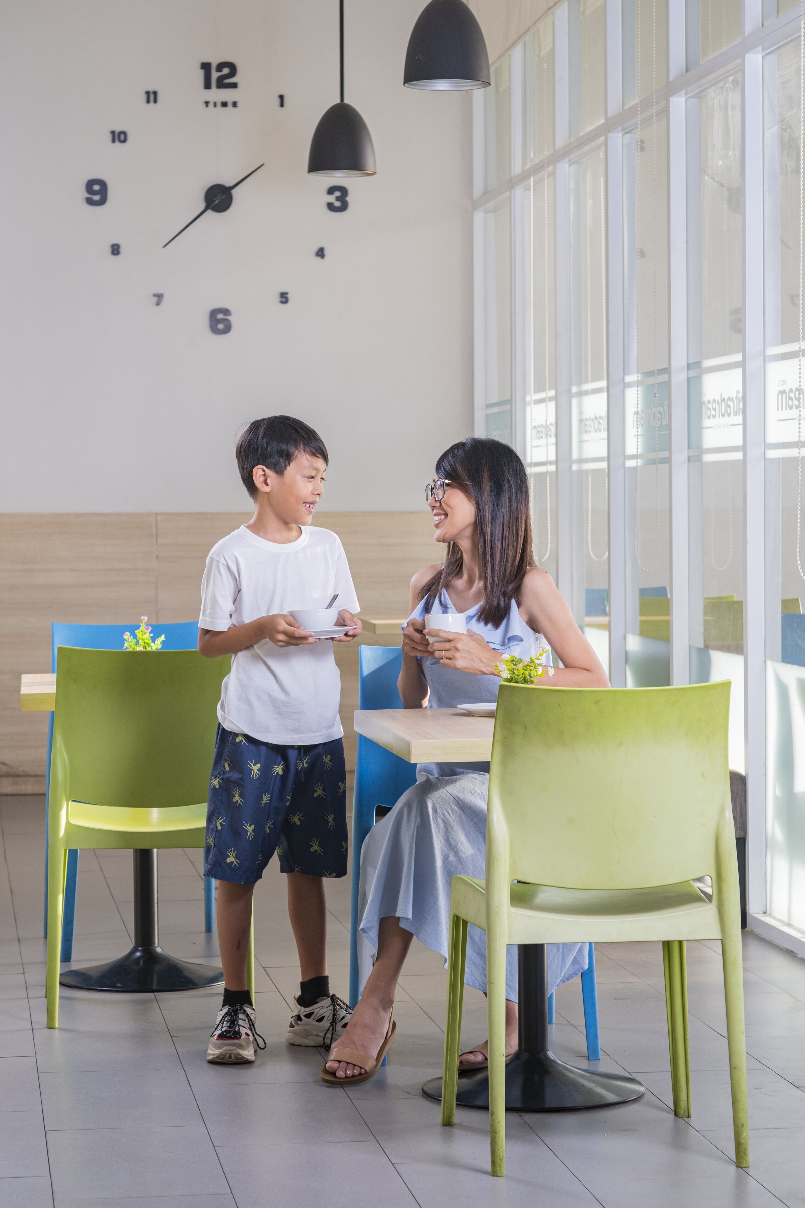 A dining area with a clock on the wall, a child and an adult seated at a table, and a window with blinds.