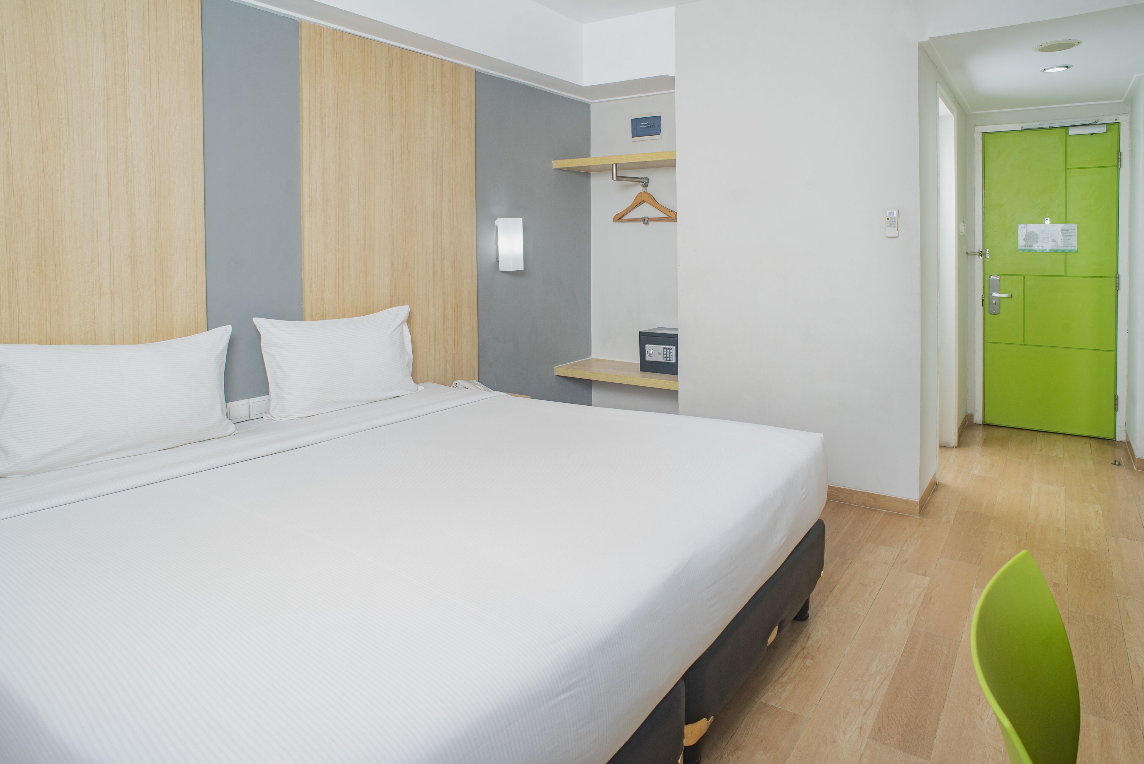 Une chambre d’hôtel avec un grand lit, un mur recouvert de panneaux de bois, une petite table avec un coffre-fort, une porte verte et une chaise verte.