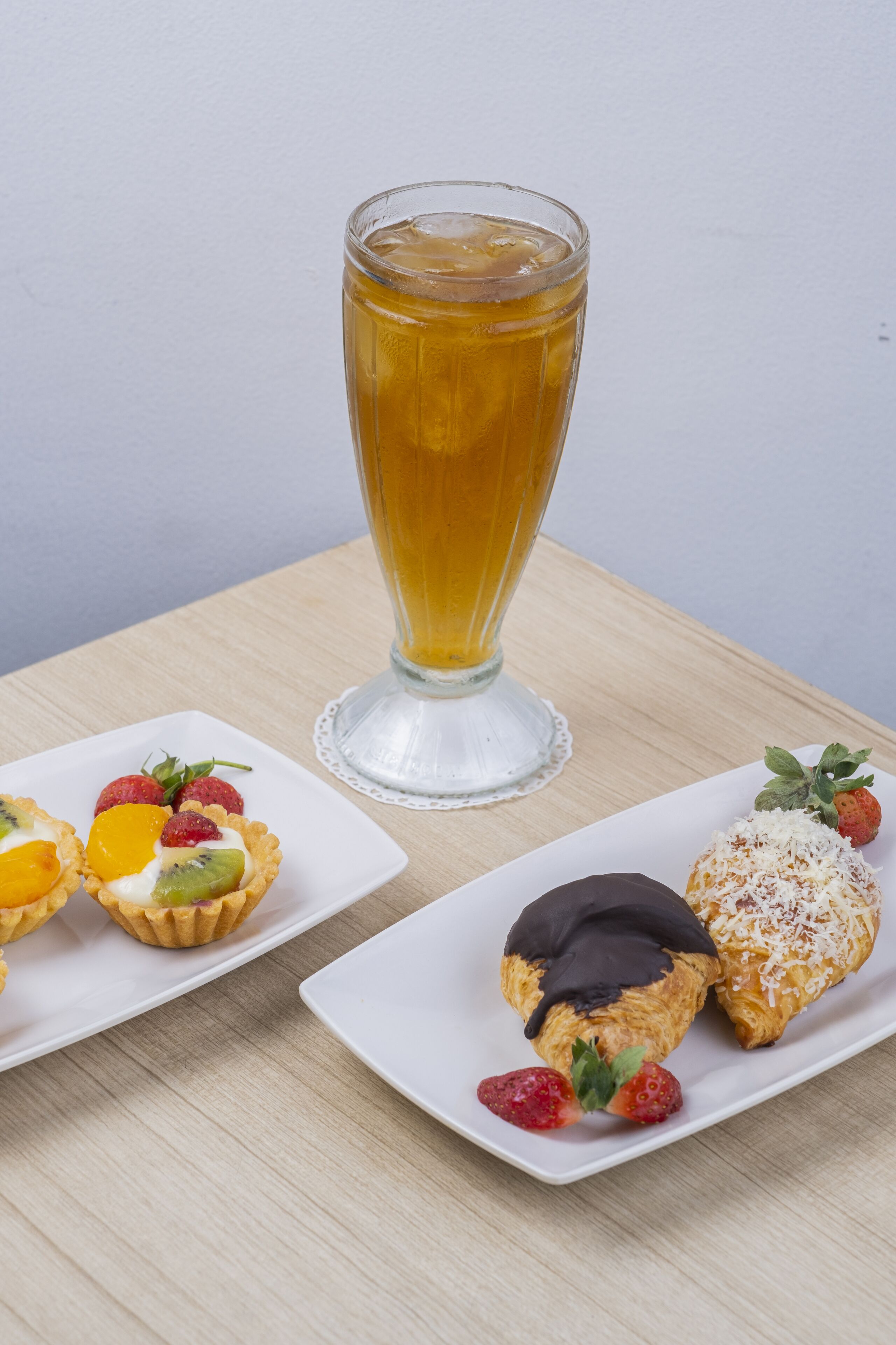 Une table avec un grand verre de thé glacé, deux assiettes carrées contenant des pâtisseries et des fruits, et un chemin de table blanc.