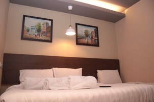 A hotel room with a bed, white linens, two framed paintings on the wall, and a hanging light fixture.