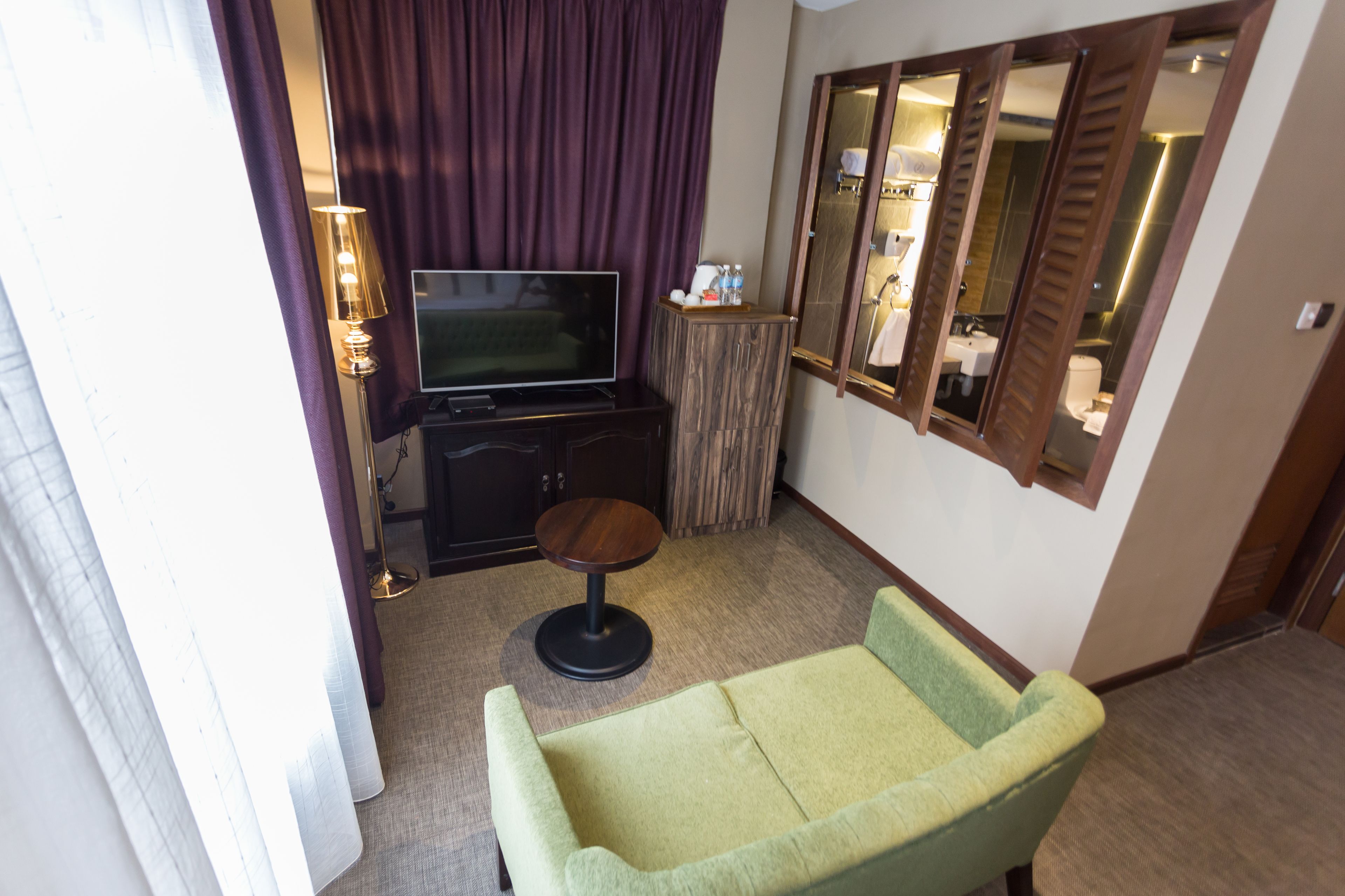A hotel room with a green sofa, a television on a wooden cabinet, a small round table, and a mirror on the wall.