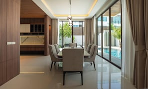 A modern dining area with a glass table and chairs, a kitchen with wooden cabinets, and a view of a swimming pool.