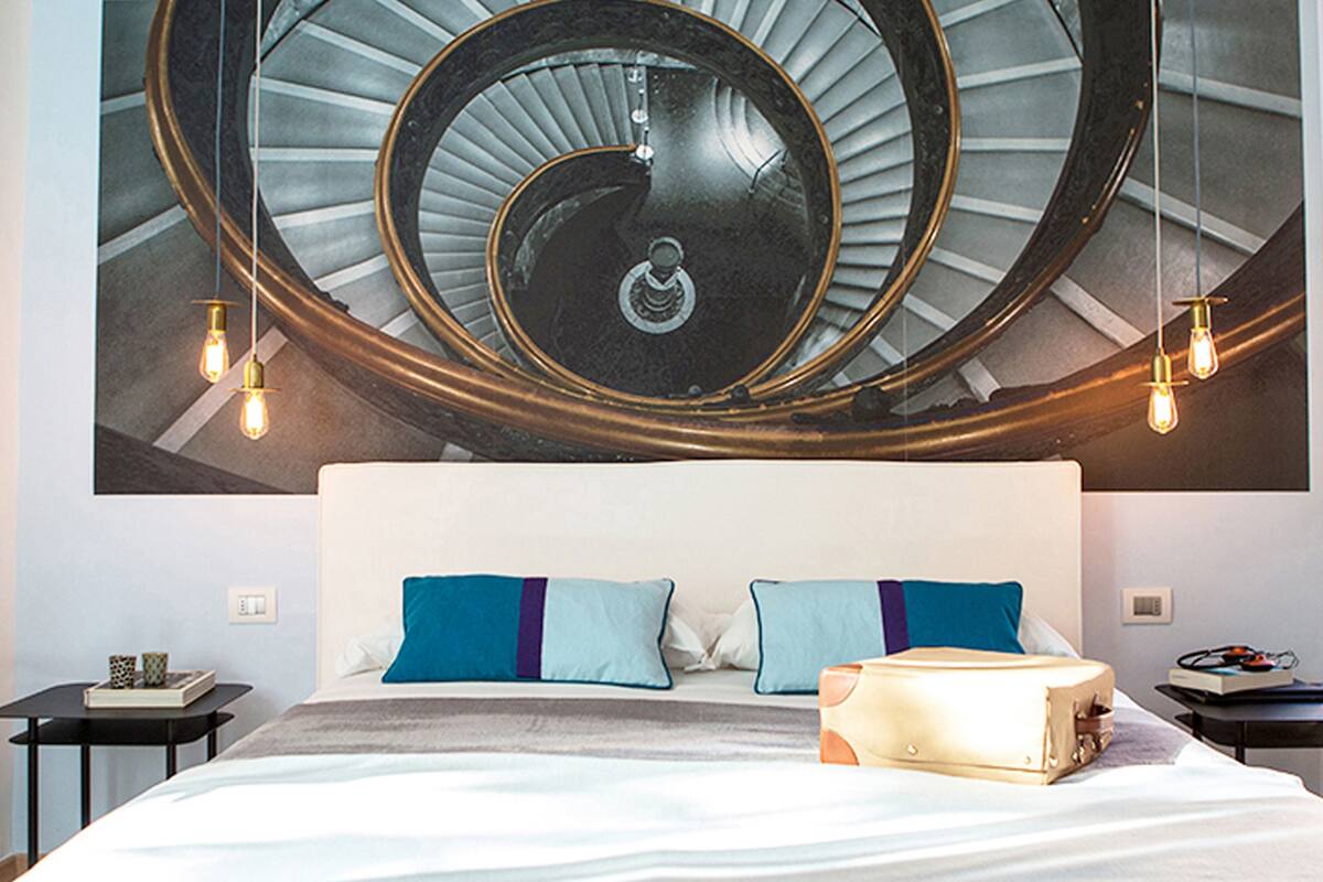 A bedroom with a large mural of a spiral staircase, a white bed with two pillows, and two bedside tables with lamps.