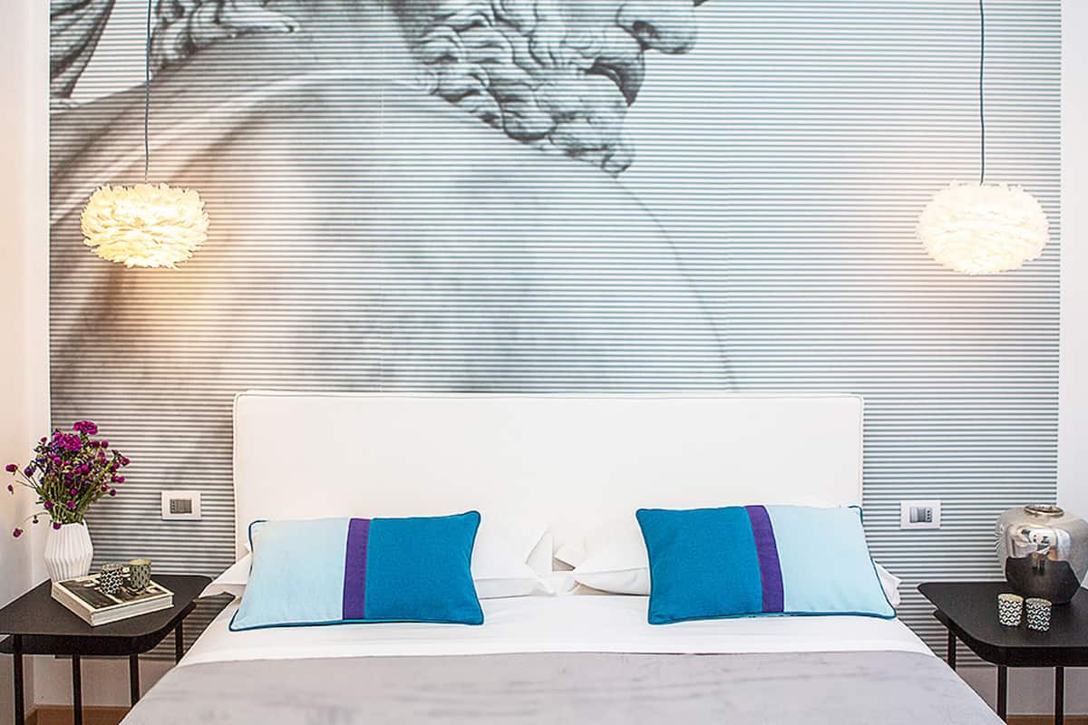 A bedroom with a large mural of a classical figure, a white bed with blue pillows, two bedside tables with decorative items, and a modern pendant light.
