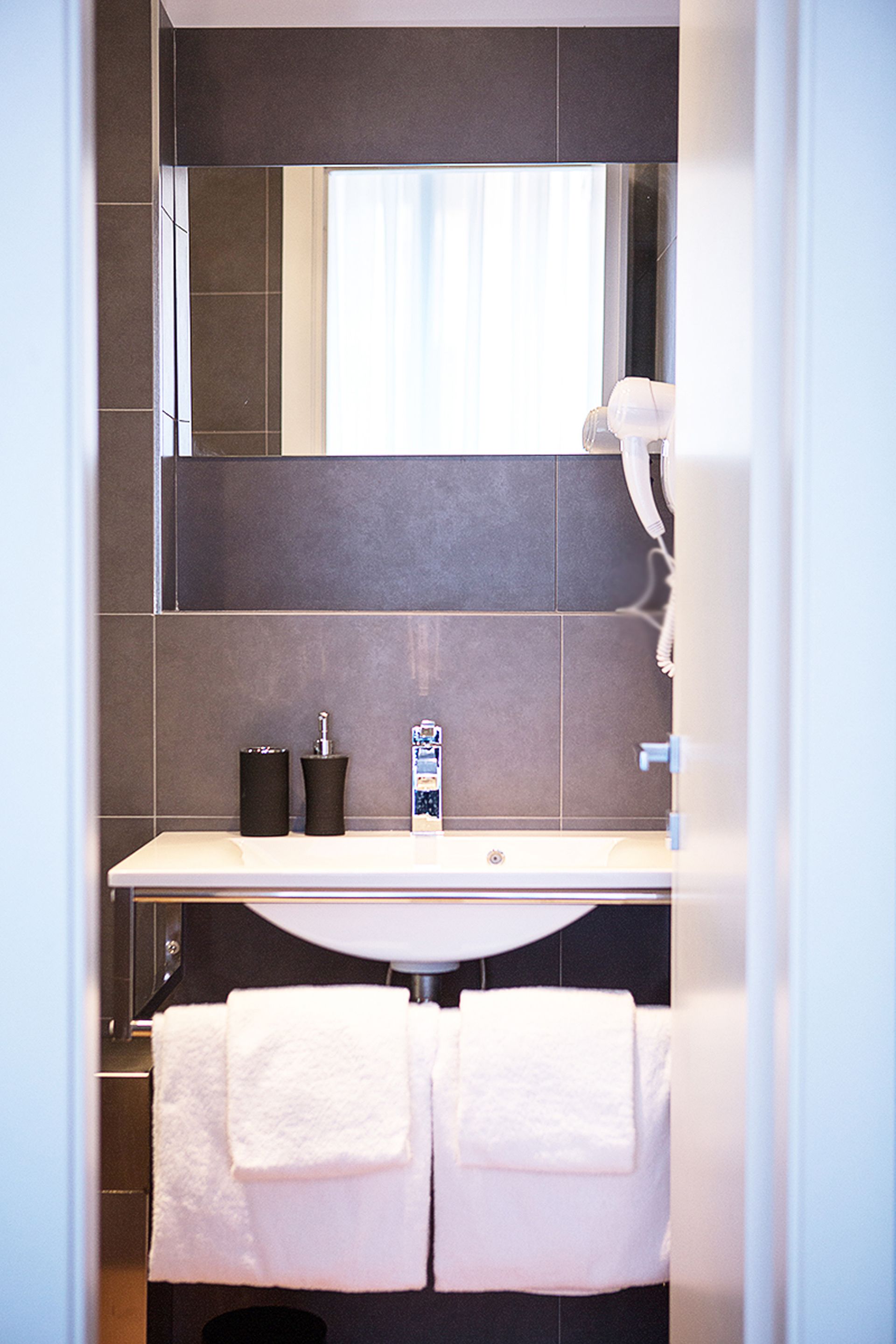 A bathroom with a white sink, dark tiles, a mirror, and two white towels hanging on a rack.