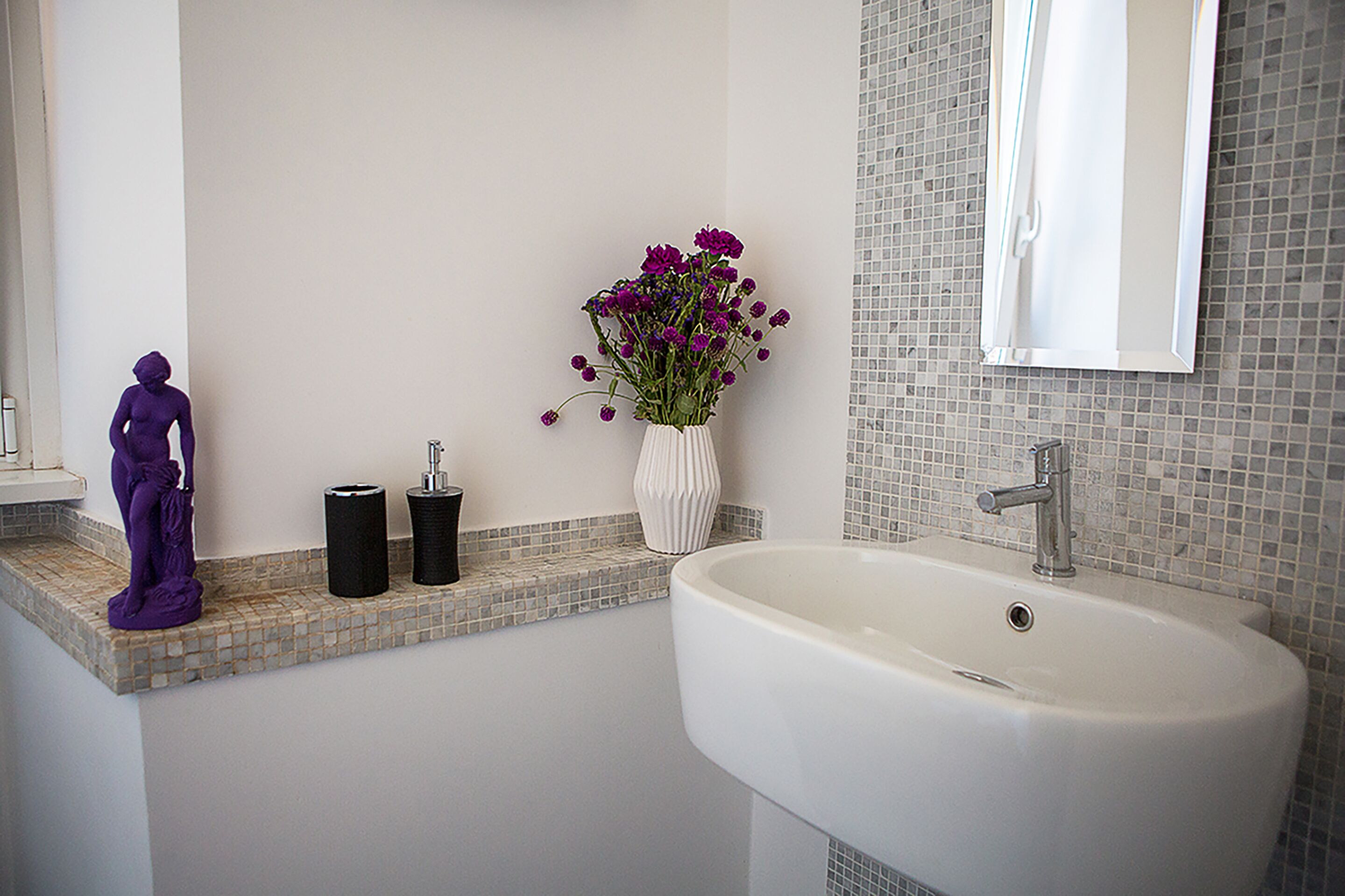A bathroom with a white sink, a mirror, a vase with purple flowers, and a statue on the counter.