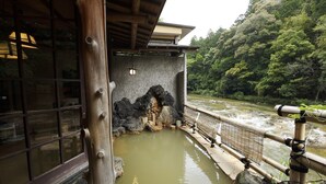 Un onsen japonés tradicional con estructura de madera, pared de piedra y vista a un río que fluye.