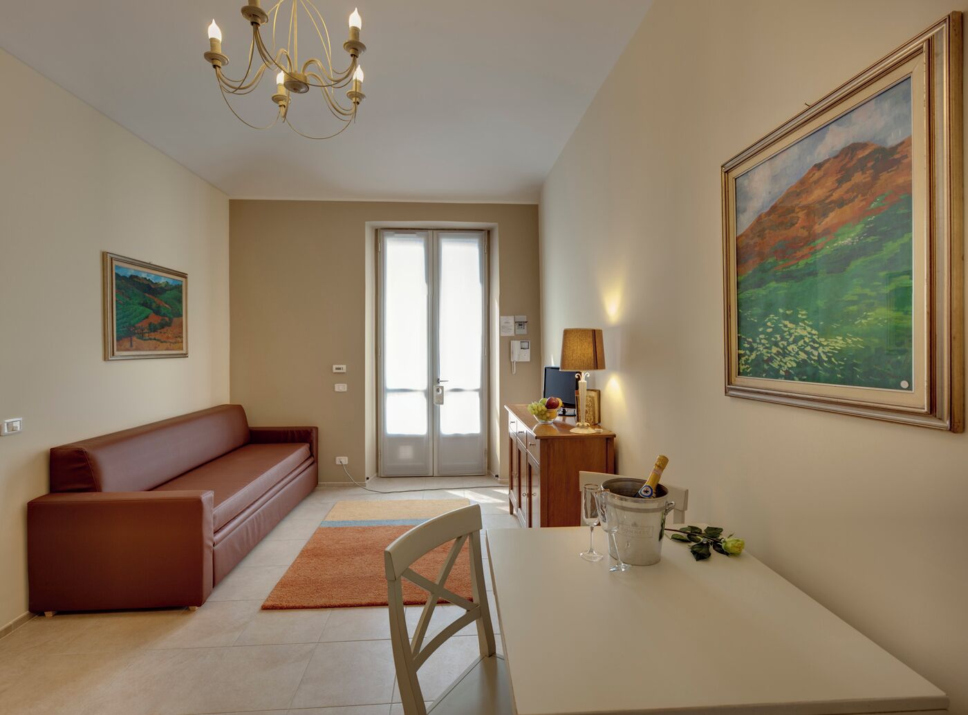Un salon comprenant un canapé, une table à manger avec une chaise, un lustre, un tableau et une porte donnant sur une autre pièce.