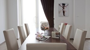 Un comedor con una mesa larga, sillas blancas, un jarrón de cristal con flores y una obra de arte enmarcada en la pared.
