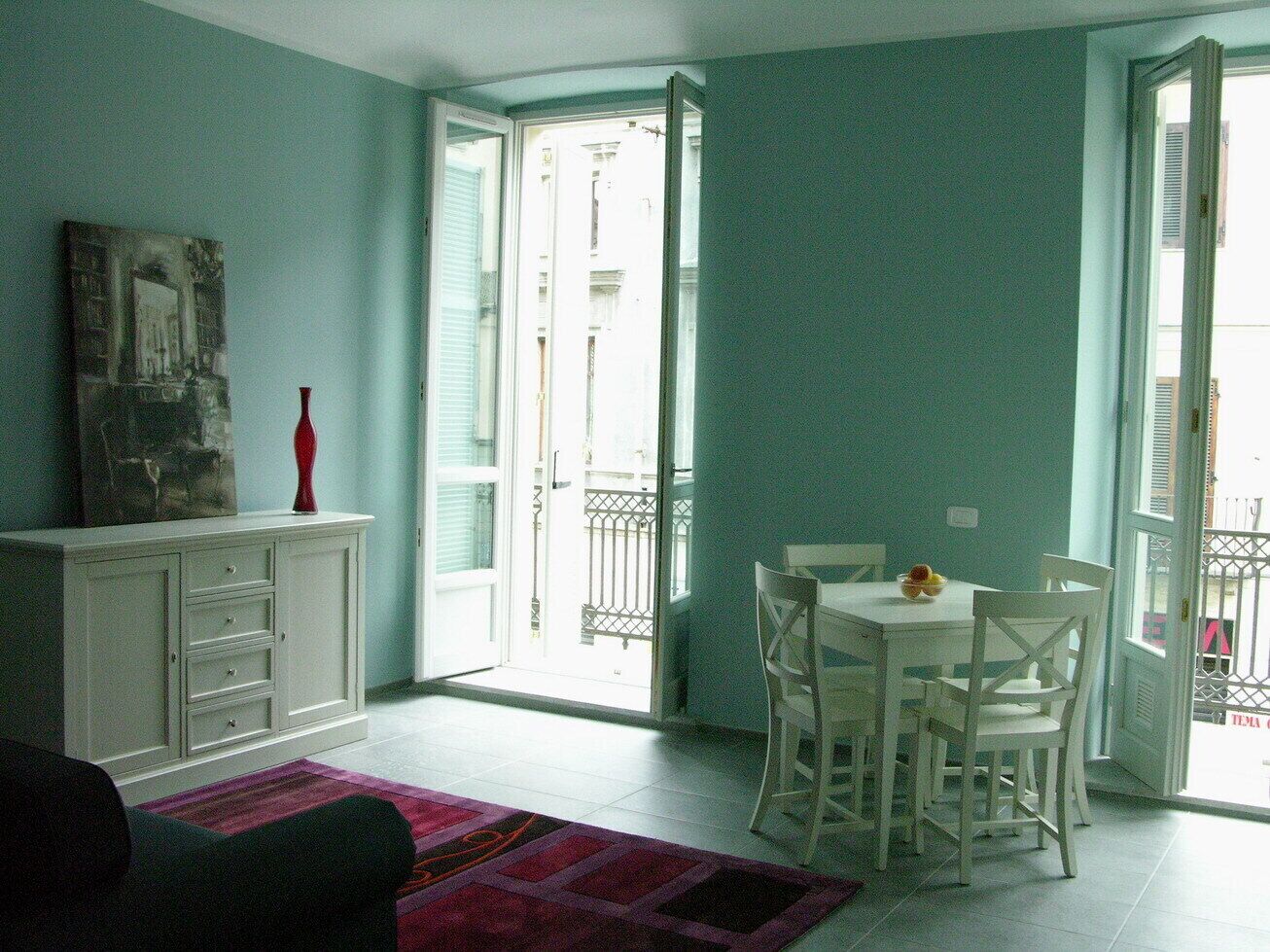 Quarto com mesa de jantar e cadeiras, um armário branco, um vaso vermelho e vista para o exterior por uma porta de vidro.