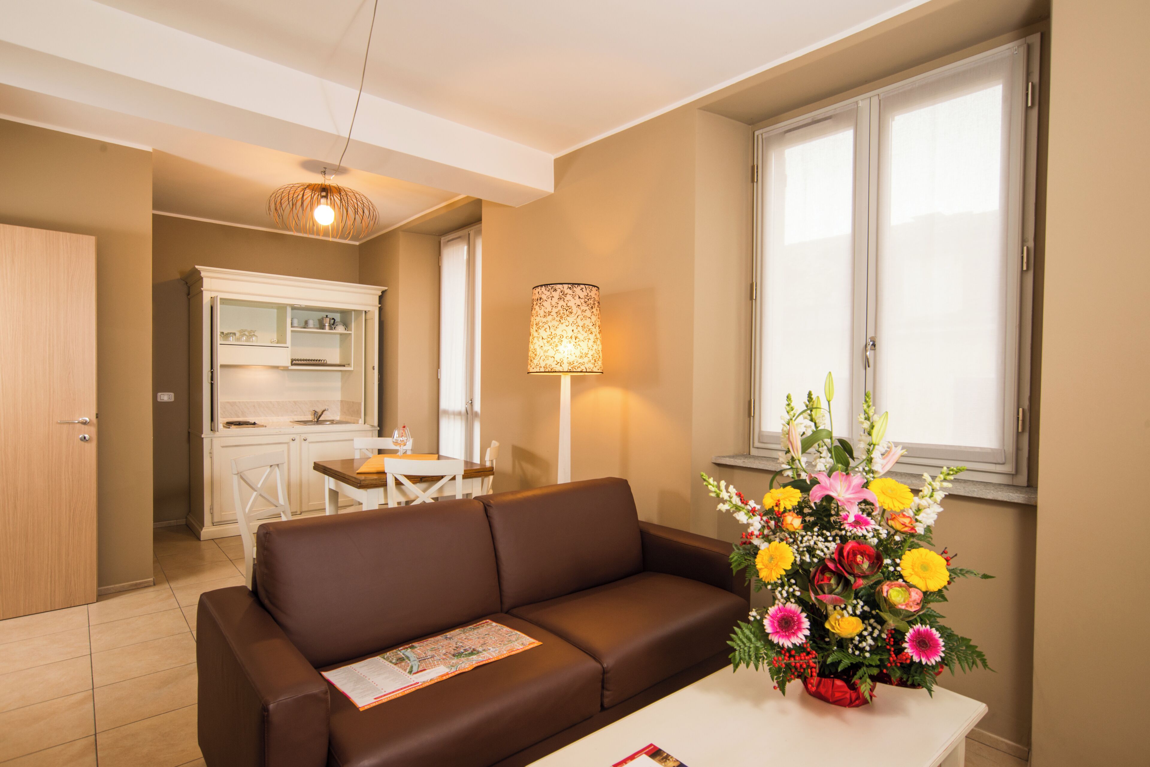Ein Wohnzimmer mit braunem Sofa, einem Esstisch, einer Küchenzeile und einem Fenster mit Blumen.