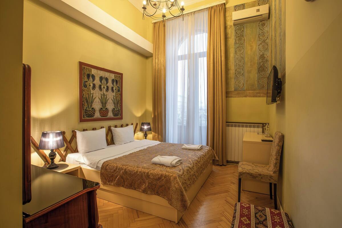 A hotel room with a bed, bedside lamps, a chair, a window with curtains, and a wall-mounted TV.