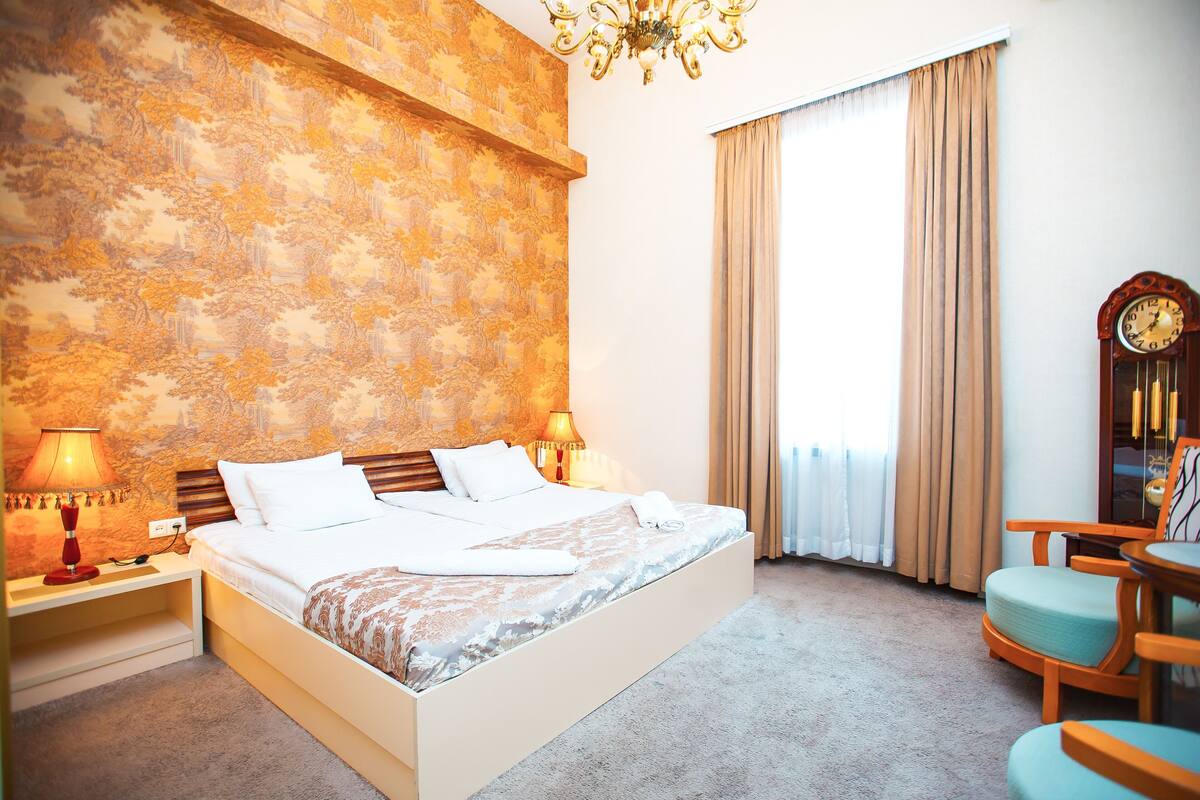 Ein Hotelzimmer mit einem großen Bett, zwei Nachttischlampen, einem Holzstuhl, einer Uhr und einem Fenster mit Vorhängen.