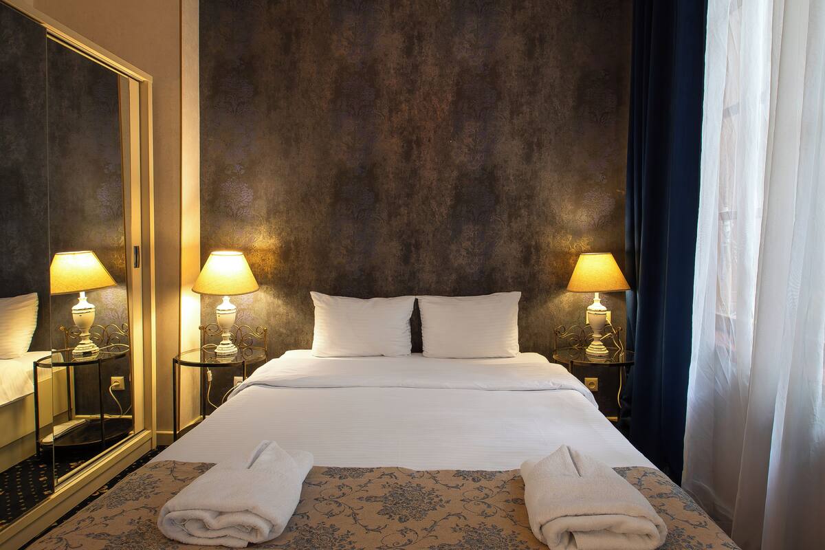 Ein Hotelzimmer mit einem großen Bett, zwei Nachttischen mit Lampen, einem Spiegel und Blick auf den Nachthimmel durch transparente Vorhänge.