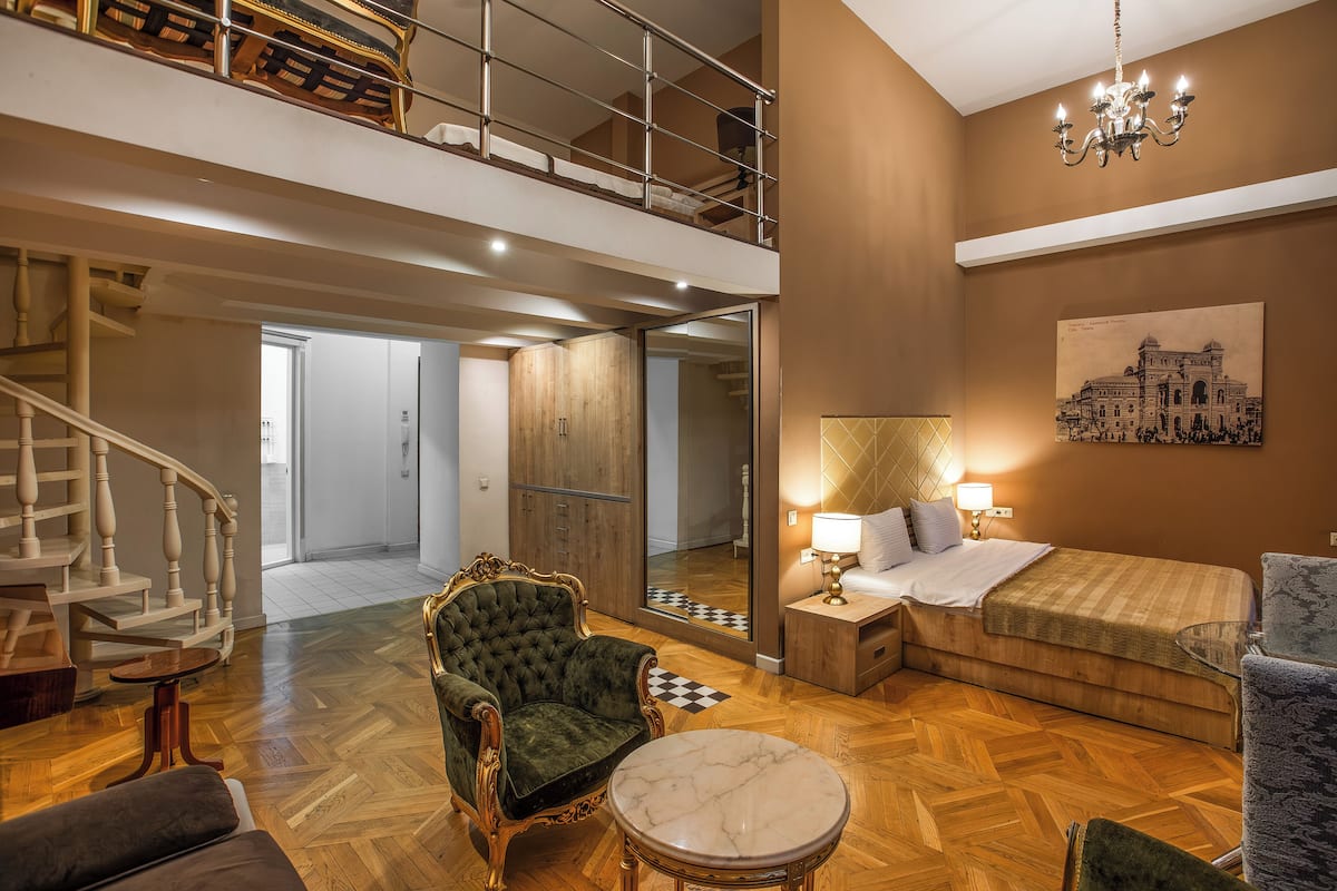 Ein geräumiges Zimmer mit einem großen Bett, einer Sitzecke mit Sessel und Tisch, einer Treppe zu einer oberen Ebene sowie einem Kronleuchter.