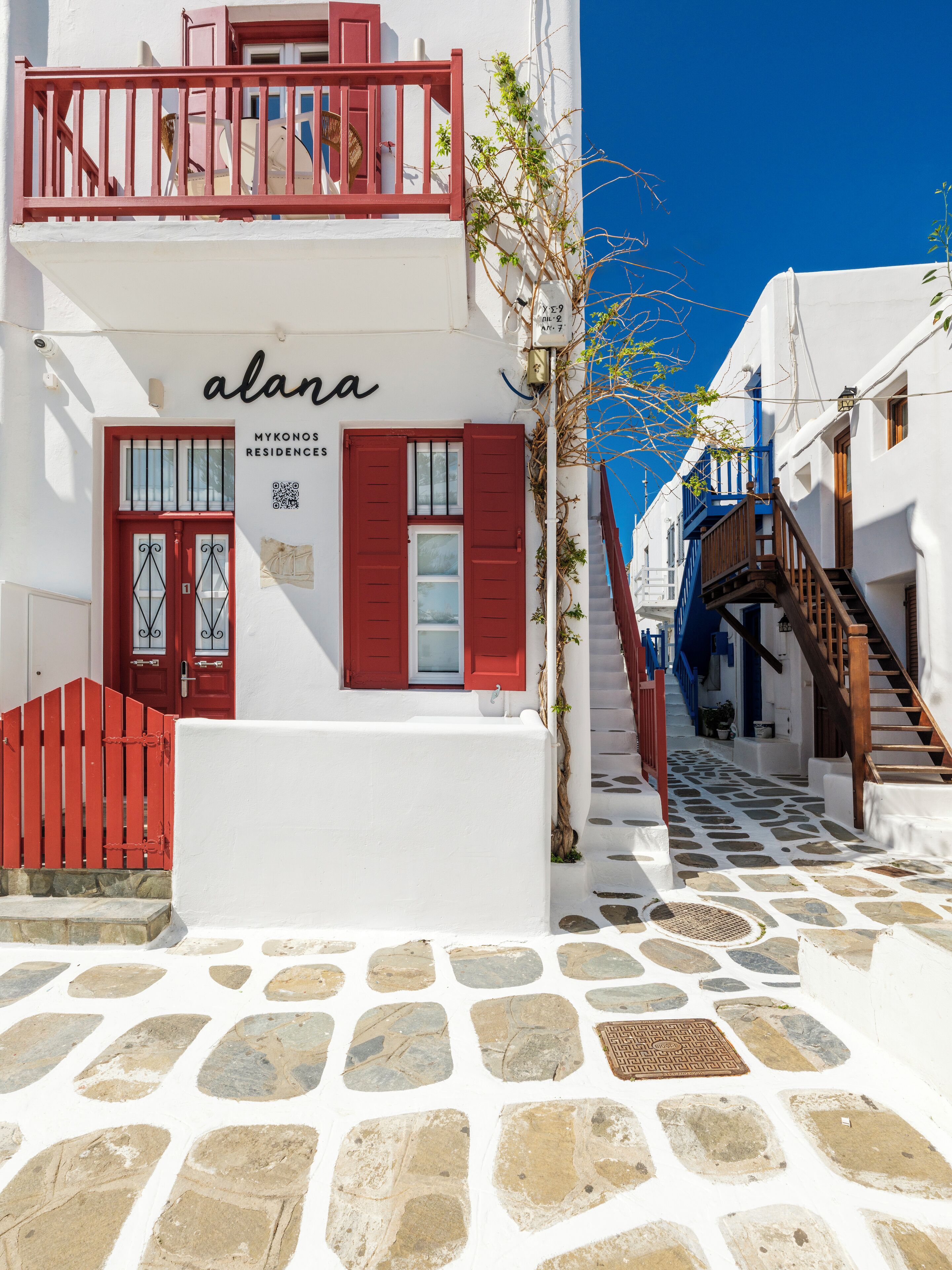 Un edificio blanco con contraventanas rojas y un balcón, con la leyenda 'alana' y 'MYKONOS RESIDENCES'.