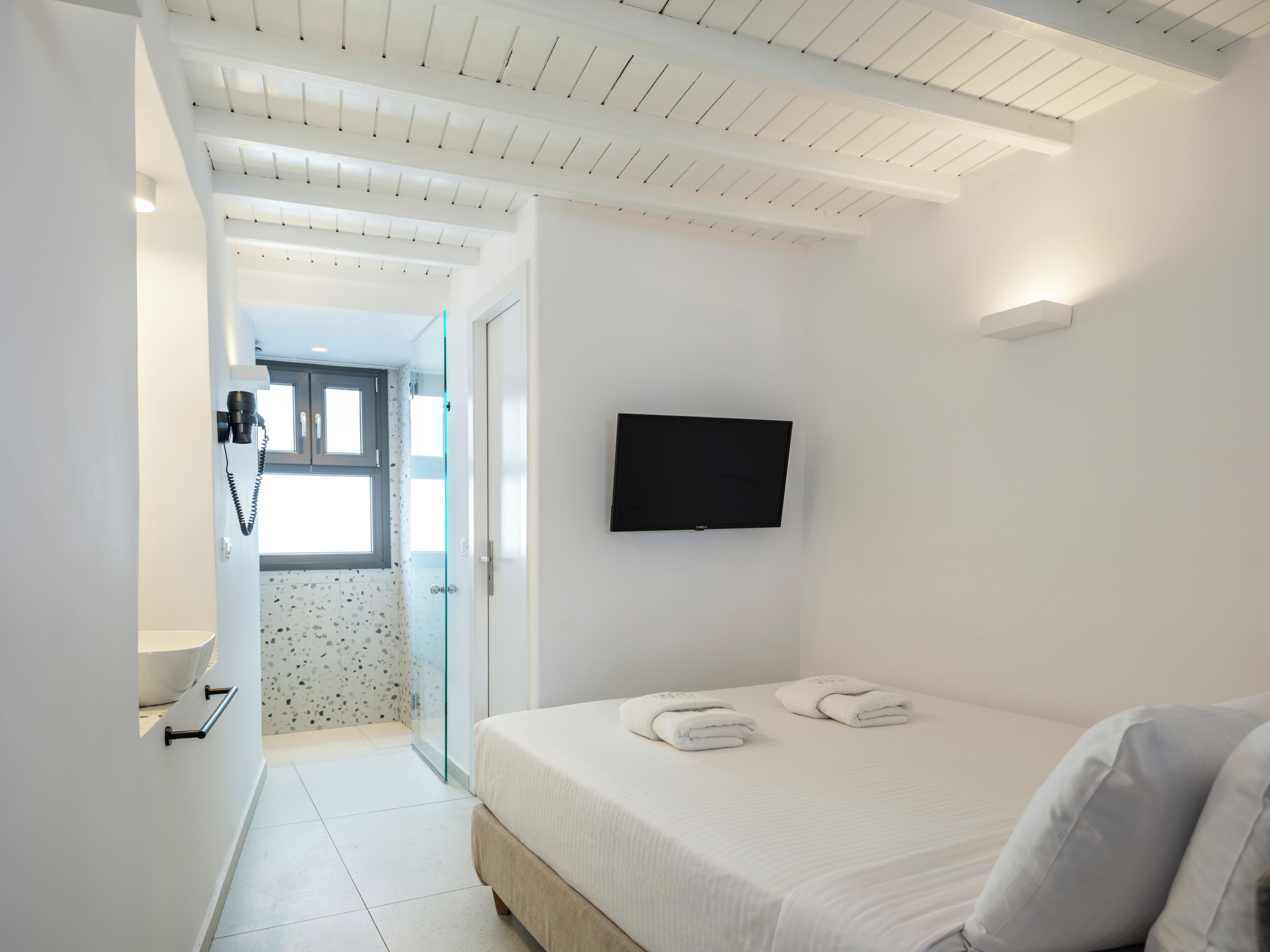 A modern hotel room with a bed, a TV mounted on the wall, and a bathroom visible through an open door.