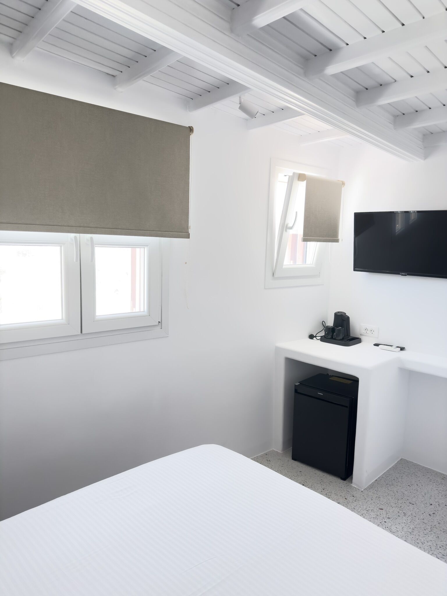 A modern bedroom with a bed, a desk with a computer and a coffee maker, a TV on the wall, and a window with a blind.